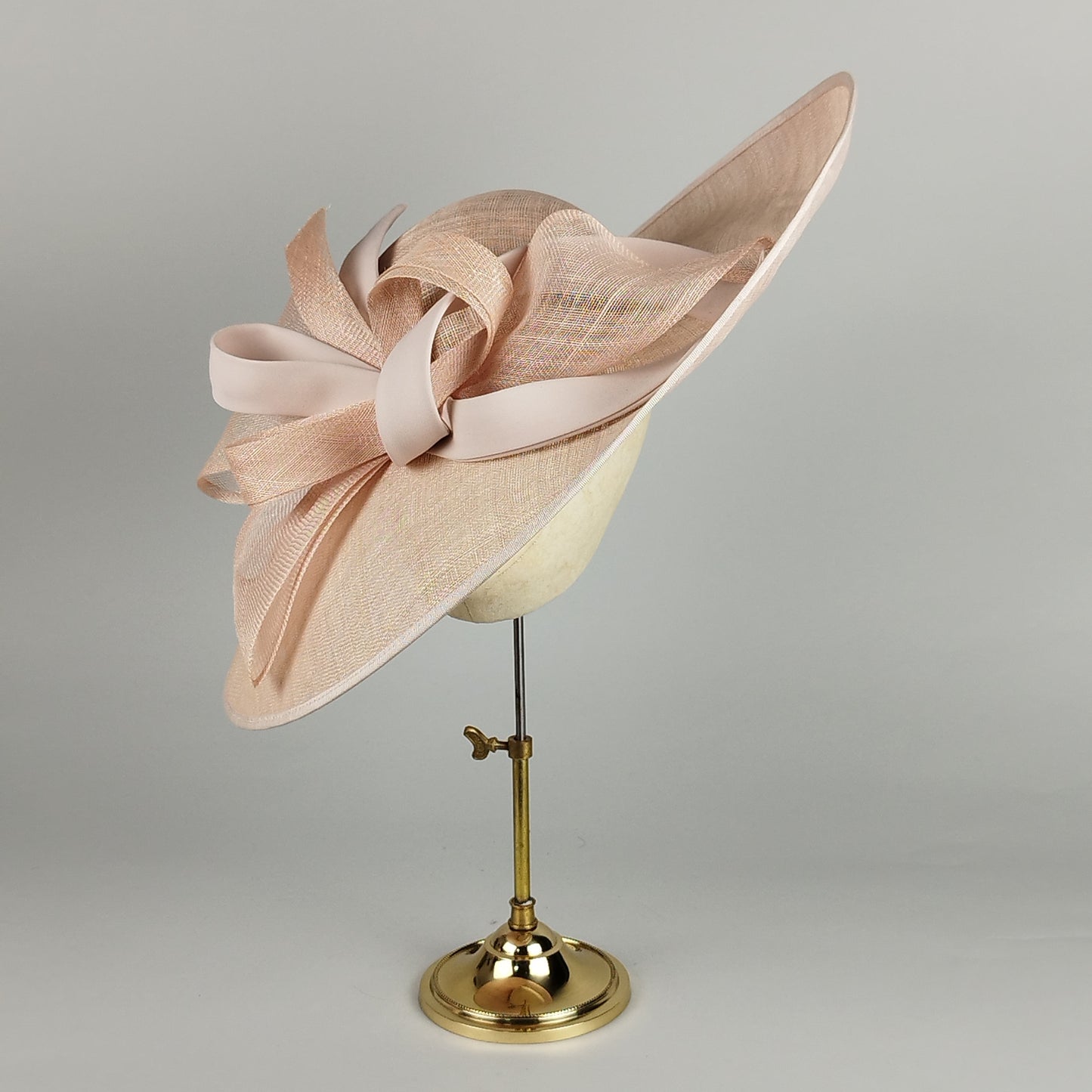 Pale pink hat with sinamay loops and swirls and a sloped brim. A sophisticated and modern design perfect for weddings, Royal Ascot, and other elegant occasions.