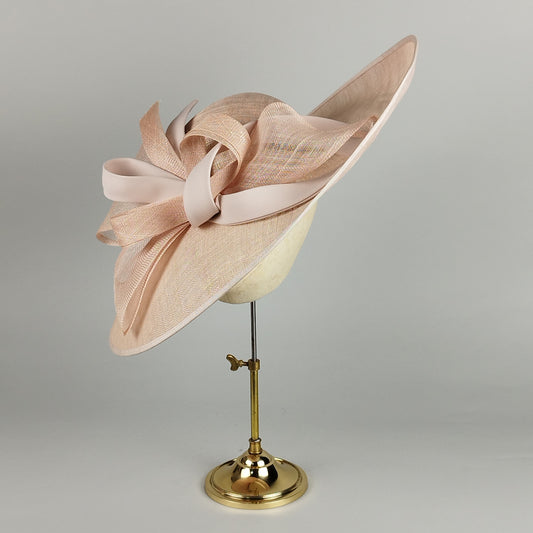 Pale pink hat with sinamay loops and swirls and a sloped brim. A sophisticated and modern design perfect for weddings, Royal Ascot, and other elegant occasions.