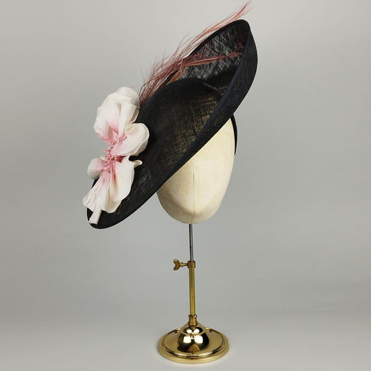 Black sinamay hatinator with large pink silk flower and feather detail, mounted on gold display stand